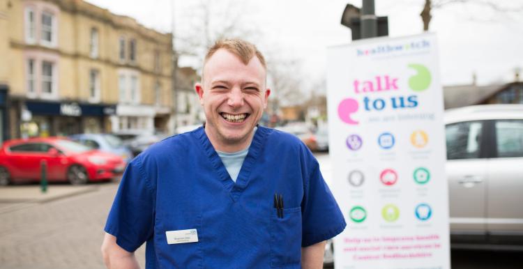 NHS worker standing infront of a Healthwatch sign that says 'talk to us'