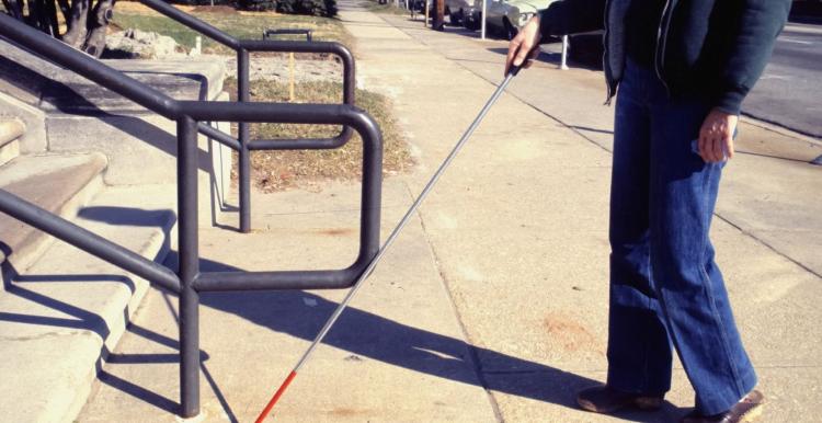 A partially-sighted person using a white stick stands on a pavement approaching some steps.