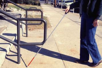 A partially-sighted person using a white stick stands on a pavement approaching some steps.